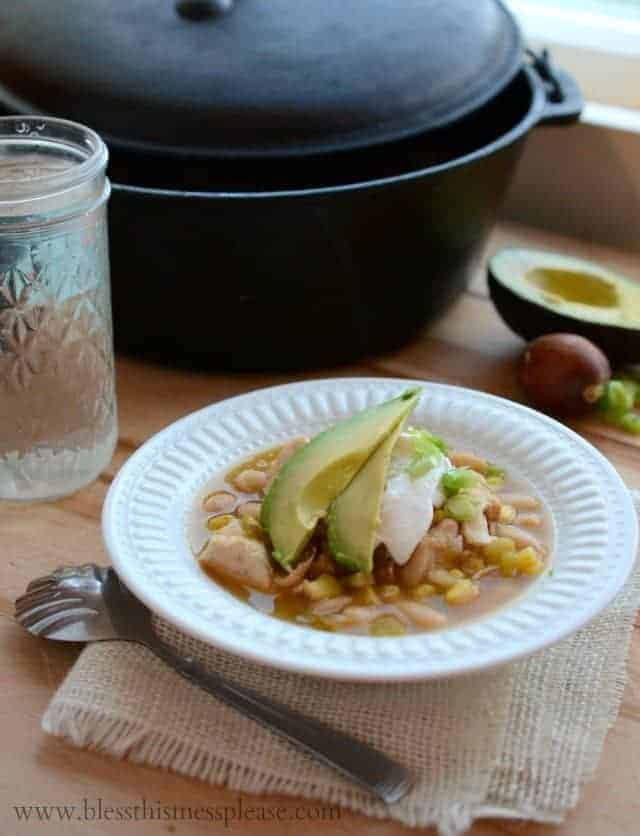 Image of a white bowl with white chicken enchilada soup garnished with two avocado slices, some sour cream, and sliced green onions. 