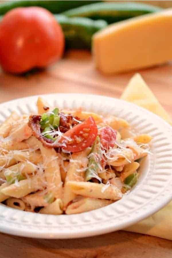 A round white bowl of penne pasta with bacon, tomato, green peppers, creamy Alfredo sauce and grated Parmesan cheese