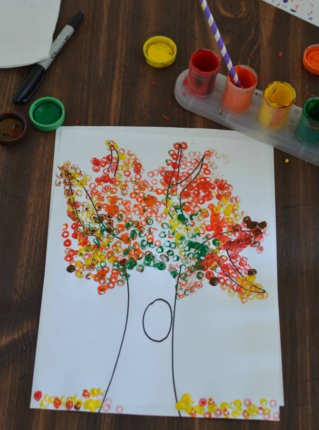 Kids fall artwork of a tree drawn on white paper with fall-colored small circles to represent leaves on the tree and ground