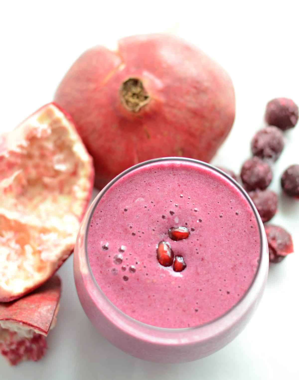 a pink smoothie in a glass cup with a pomegranate and pomegranate seeds to the side