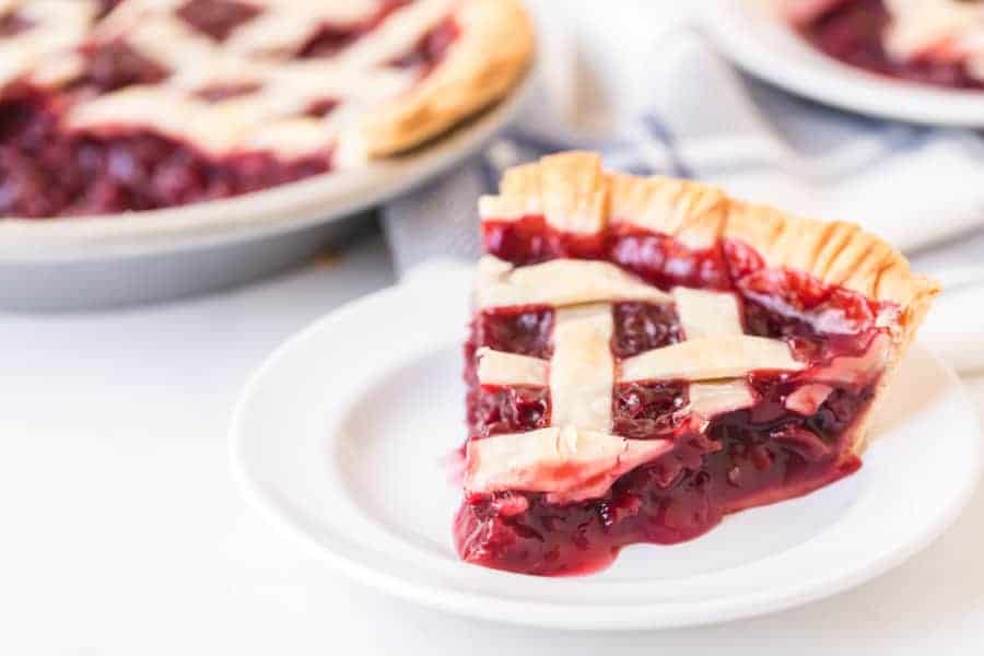 cherry pie slice on round white plate next to cherry pie in metal pie plate on white surface