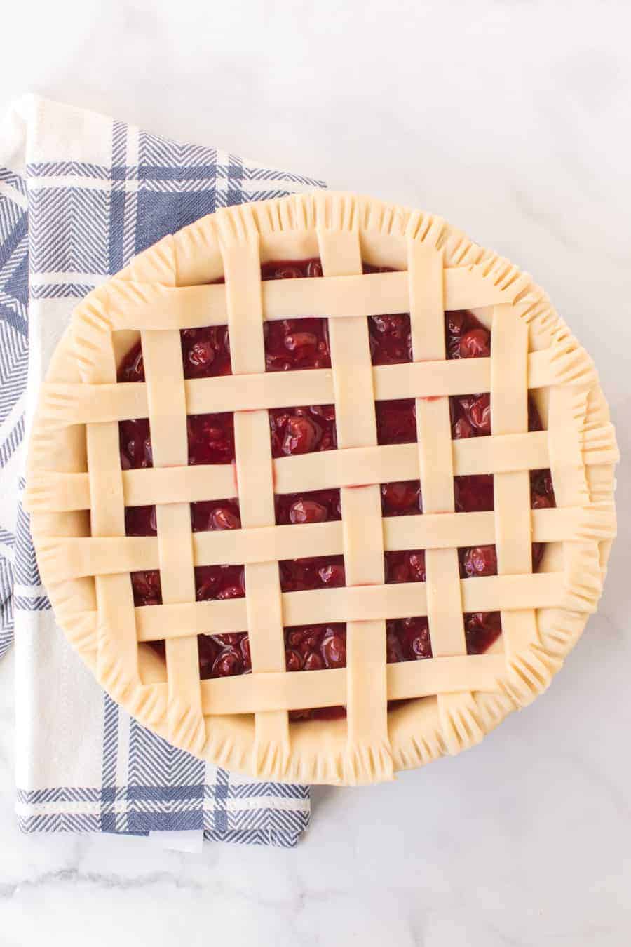 unbaked cherry pie with criss cross pie crust resting on blue and white checkered towel on white countertop