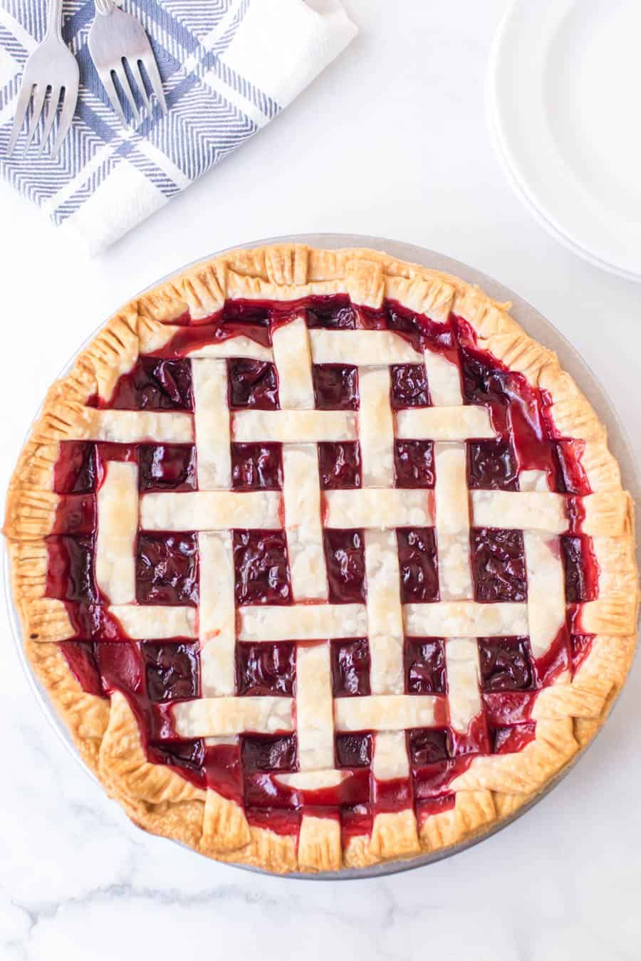 baked cherry pie with criss cross pie crust resting on white countertop next to two forks with blue and white checkered towel