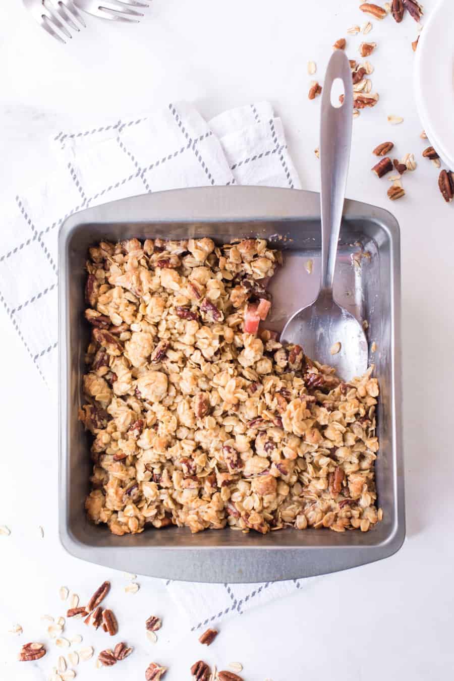 rhubarb crisp in baking pan with serving spoon on white towel and white countertop with pecans