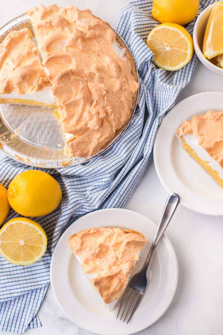 slice of lemon meringue pie on round white plate next to lemons andpie tin