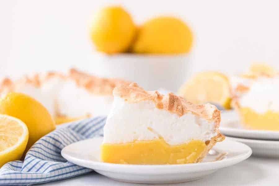 slice of lemon meringue pie on white plate with blue checkered towel and lemons