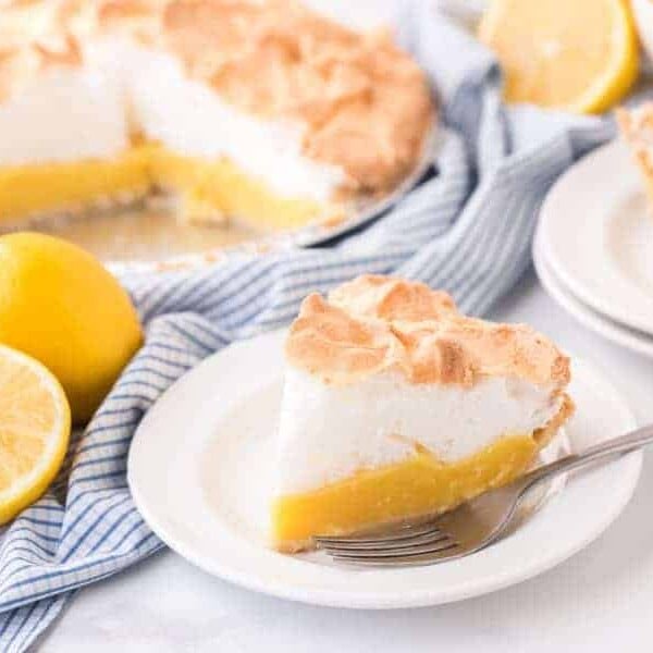closeup of lemon meringue pie on white plate with fork next to lemons and pie