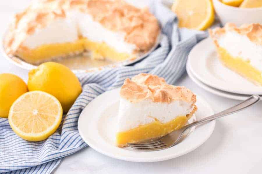 slice of lemon meringue pie on white plate with fork next to lemons and blue checkered towel