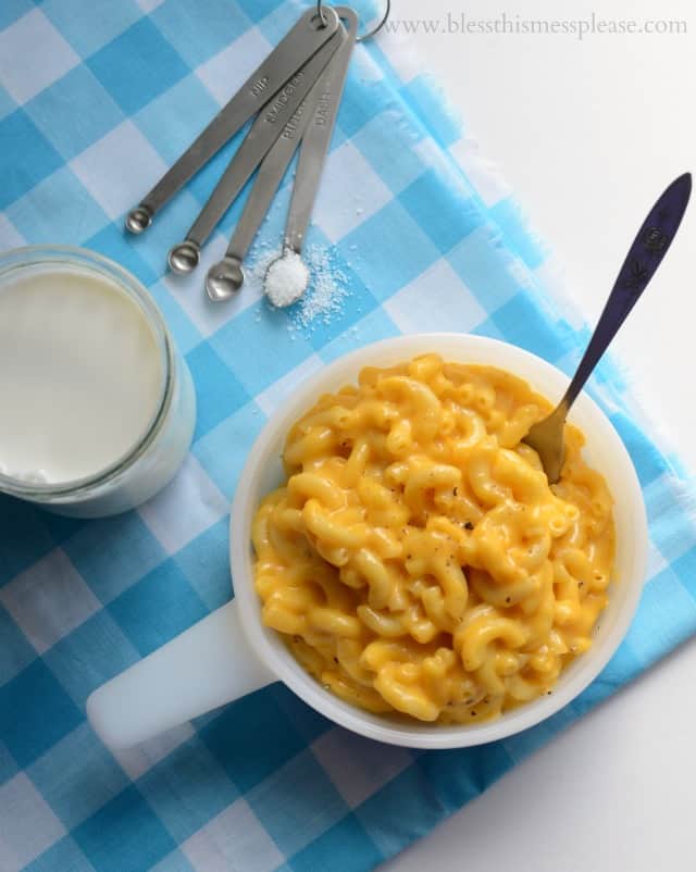 White bowl with yellow macaroni and cheese on a blue and white cloth with fork in bowl and measuring spoon with white salt and glass with white milk.