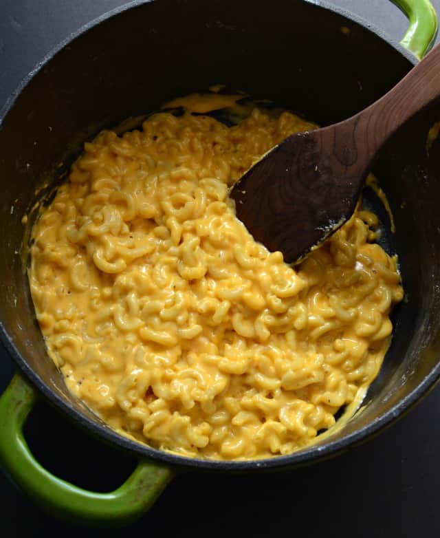 Black pot with green handles, with yellow macaroni and cheese and wooden spoon inside.