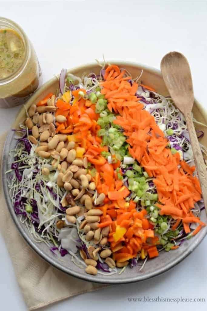 A serving bowl of chopped Asian peanut salad with rows of shredded cabbage, peanuts, chopped bell peppers, scallions, and shredded carrots