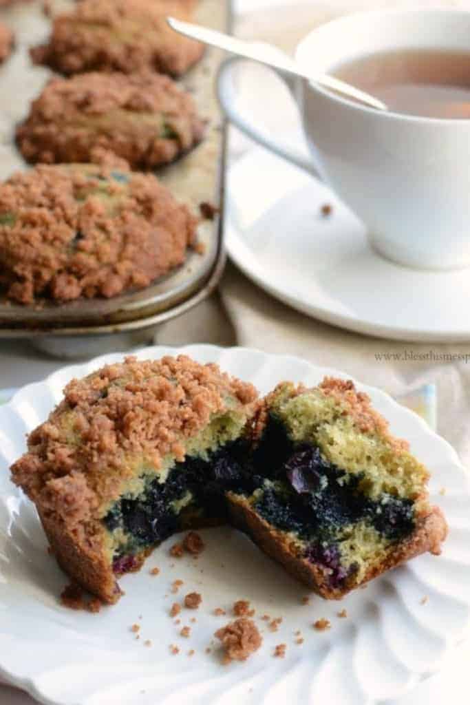 A blueberry muffin with crumb topping on a white plate with a cup of tea and a muffin tin of muffins in the background