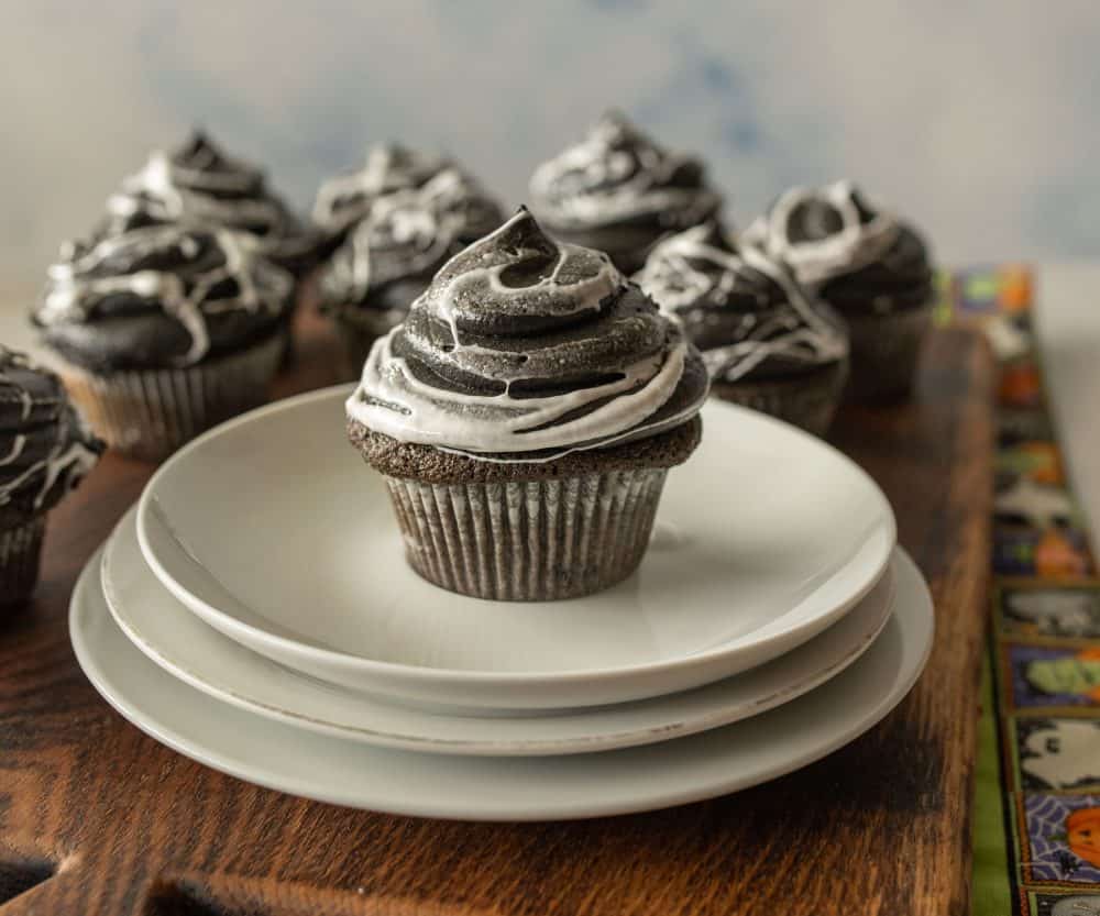 one spiderweb cupcake on a stack of white plates with a few other cupcakes in the background
