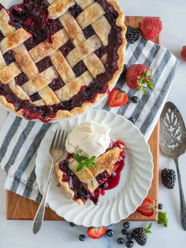 top view of a slice of triple berry pie on a plate next to the pie, serving utensils, and berries