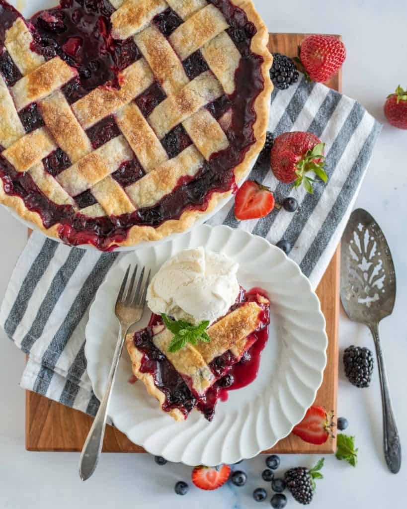 top view of a slice of triple berry pie on a plate next to the pie, serving utensils, and berries