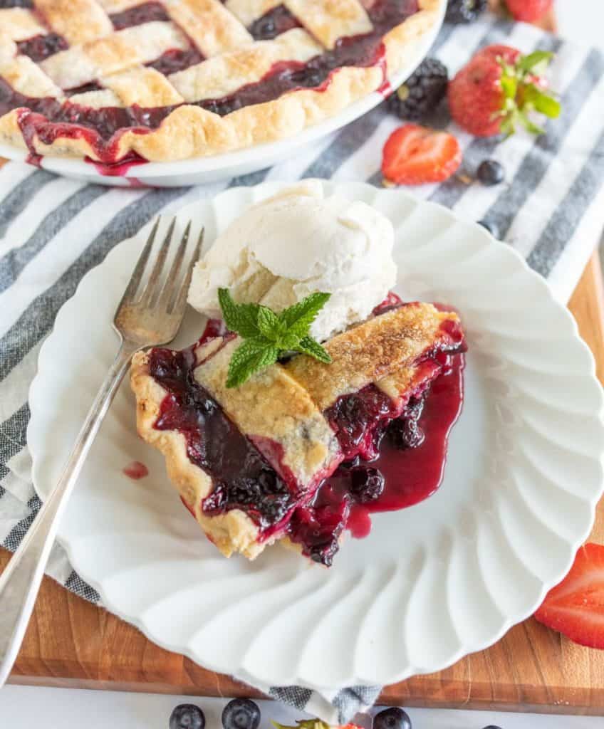close up picture of a slice of triple berry pie on a plate with ice cream and a mint leaf