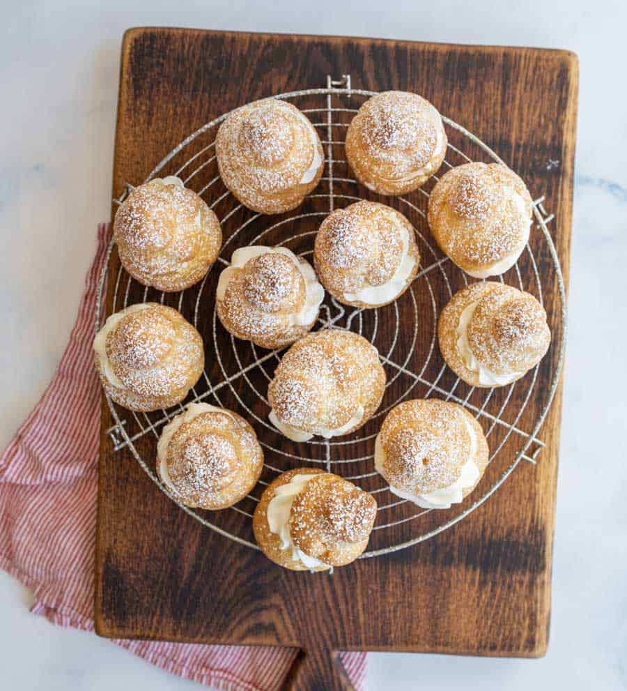 Top view of cream puffs dusted with powdered sugar on a cooling rack on a cutting board.