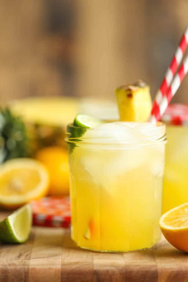 A glass of pineapple lemonade with two straws, fresh pineapple and a lime wedge
