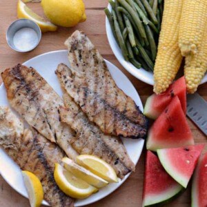 A plate with three grilling fish fillets and lemon wedges is surrounded by fresh green beans, corn on the cob, watermelon slices, a whole lemon, and a small dish of salt on a wooden surface.
