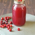 A jar of homemade juice with cherries on top sits on a round gray mat, showcasing a delightful cherry juice recipe. A small cluster of fresh cherries is scattered around the jar against a dark wooden background.