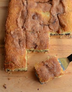 A cinnamon coffee cake with a golden-brown crust, reminiscent of a delectable blueberry boy bait, rests on a wooden surface. A square piece has been cut and is being lifted with a spatula, revealing its moist texture and sugar-cinnamon topping.