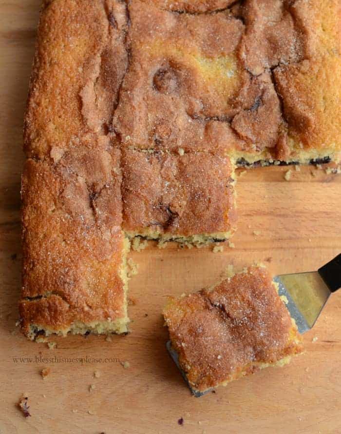 A cinnamon coffee cake with a golden-brown crust, reminiscent of a delectable blueberry boy bait, rests on a wooden surface. A square piece has been cut and is being lifted with a spatula, revealing its moist texture and sugar-cinnamon topping.
