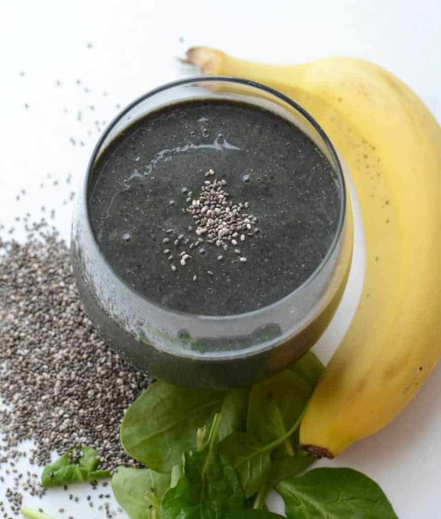 a dark colored smoothie in a glass with a banana and greens to the side