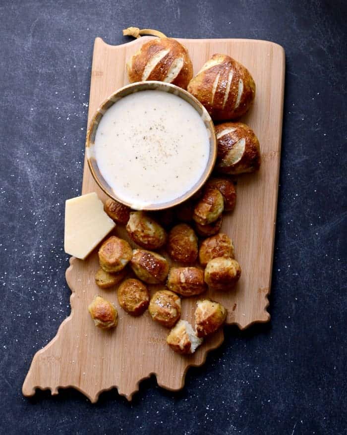 Baked parmesan pretzel bites on a bamboo cutting board in the shape of the state of Indiana with a bowl of the parmesan dipping sauce sitting on a dark blue countertop. 