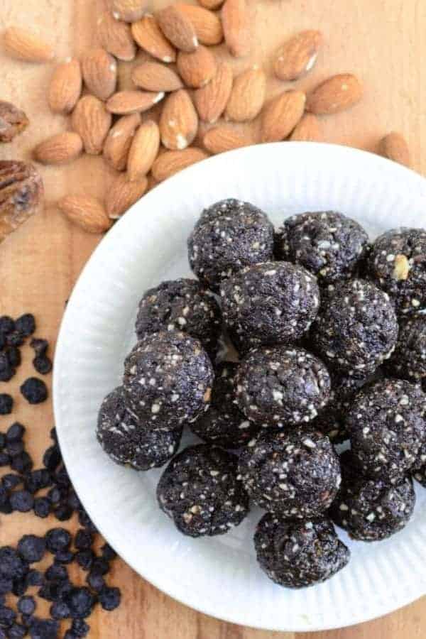 A wooden cutting board with dried blueberries and almonds and a white plate with a pile of blueberry muffin date balls