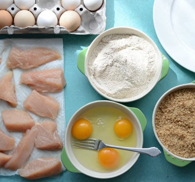 ingredients for homemade chicken nuggets on a blue background - eggs, raw chicken pieces, eggs, flour, breadcrumbs in green bowls.