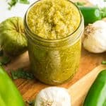 A jar of green sauce from a green sauce recipe for enchiladas sits on a wooden board surrounded by ingredients: garlic cloves, green peppers, and tomatillos. Fresh cilantro is scattered around, adding a pop of color.