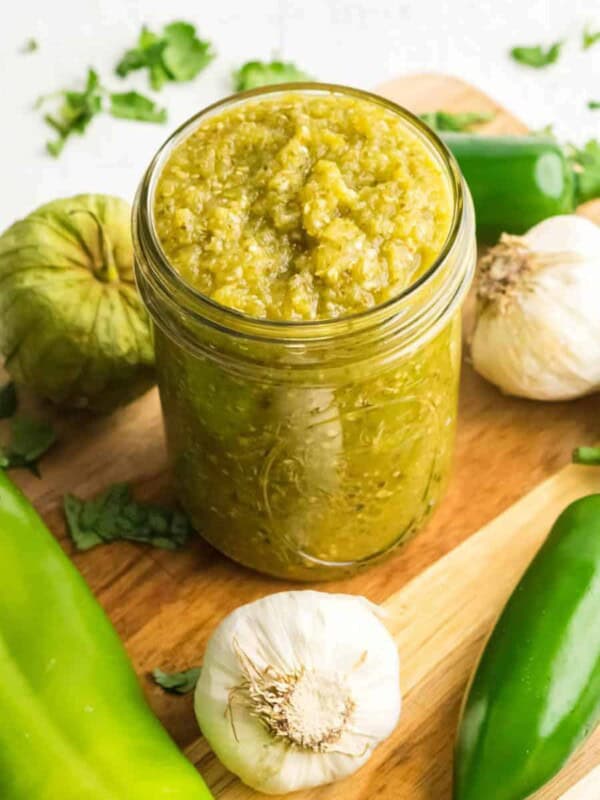 A jar of green sauce from a green sauce recipe for enchiladas sits on a wooden board surrounded by ingredients: garlic cloves, green peppers, and tomatillos. Fresh cilantro is scattered around, adding a pop of color.