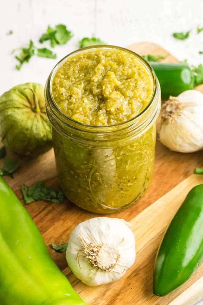 A jar of green sauce from a green sauce recipe for enchiladas sits on a wooden board surrounded by ingredients: garlic cloves, green peppers, and tomatillos. Fresh cilantro is scattered around, adding a pop of color.