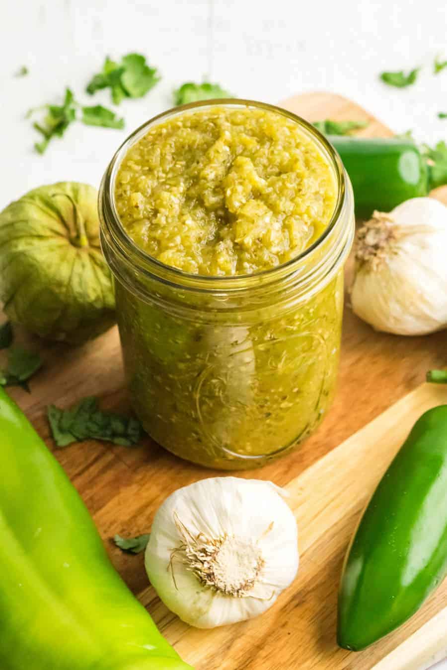 A jar of green sauce from a green sauce recipe for enchiladas sits on a wooden board surrounded by ingredients: garlic cloves, green peppers, and tomatillos. Fresh cilantro is scattered around, adding a pop of color.