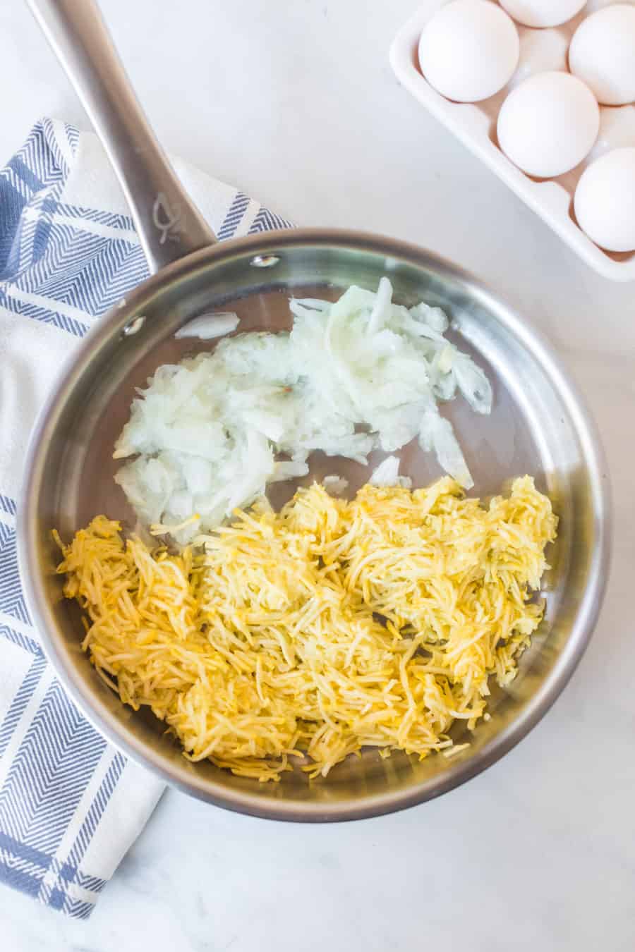 A metal pan on a marble surface holds shredded yellow potatoes and grated white onions, perfect for starting an egg bake. Nearby are a carton of eggs and a blue-striped kitchen towel.