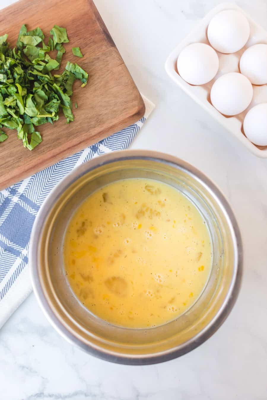 A metal bowl of beaten eggs sits on a white countertop next to a blue-and-white towel, a wooden cutting board with chopped greens for an egg bake, and a carton holding five white eggs.