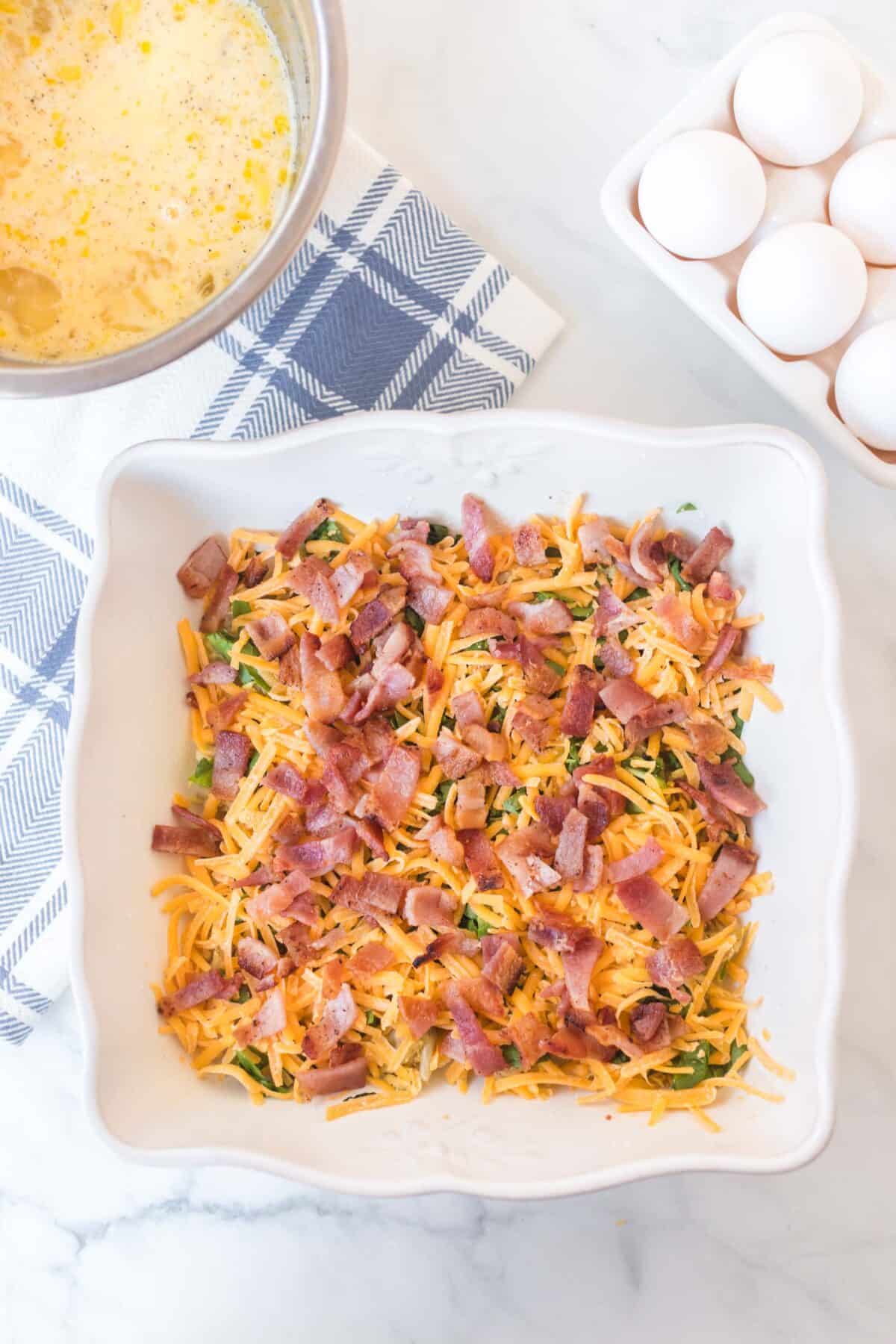 A white baking dish with chopped bacon, shredded cheese, and greens sits on a marble surface, ready for an egg bake. Nearby are a bowl of whisked eggs, a blue-and-white towel, and a carton of eggs.