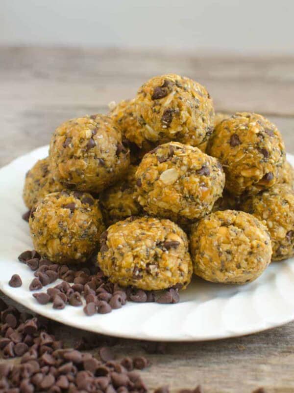 A plate of homemade pumpkin energy balls stacked in a pyramid on a wooden surface. They boast visible oats and chocolate chips, with scattered chocolate chips encircling the plate.
