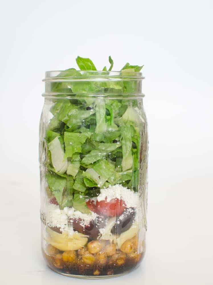 A mason jar filled with layered salad ingredients, including chickpeas and dressing at the bottom, grape tomatoes, olives, cheese, and topped with chopped lettuce—perfect for pairing with a bowl of chicken noodle soup. The background is plain white.