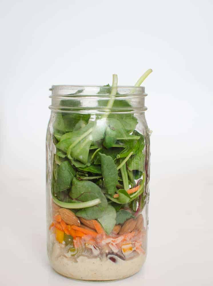 A mason jar filled with layered salad ingredients—salad dressing at the bottom, then beans, chopped vegetables, almonds, shredded carrots, and fresh spinach leaves on top—offers a refreshing twist on classic chicken noodle soup flavors.