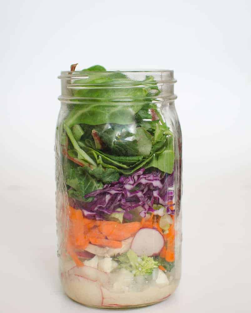 A glass mason jar filled with layers of fresh vegetables—radishes, carrots, chopped cabbage, and leafy greens—evokes the vibrant colors you might find in a hearty chicken noodle soup, all arranged neatly against a plain white background.
