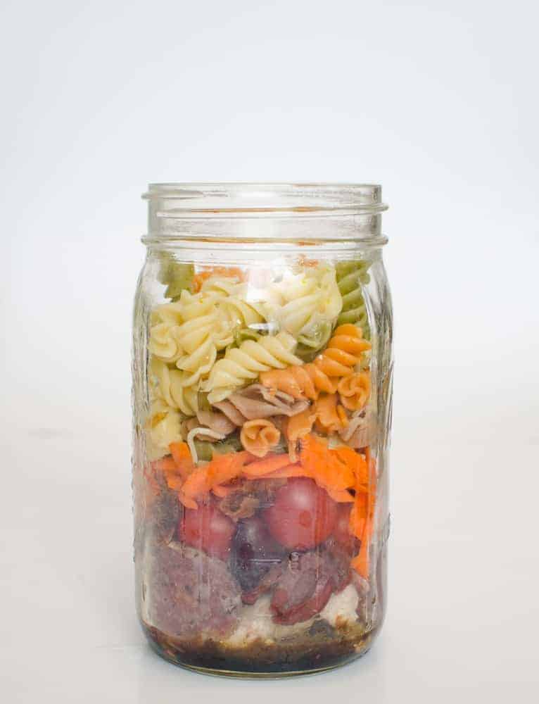 A mason jar filled with layers of ingredients, including vinaigrette, white beans, red grapes, shredded carrots, shredded chicken—bringing a twist on chicken noodle soup—and tri-color rotini pasta, all stacked visibly from bottom to top.
