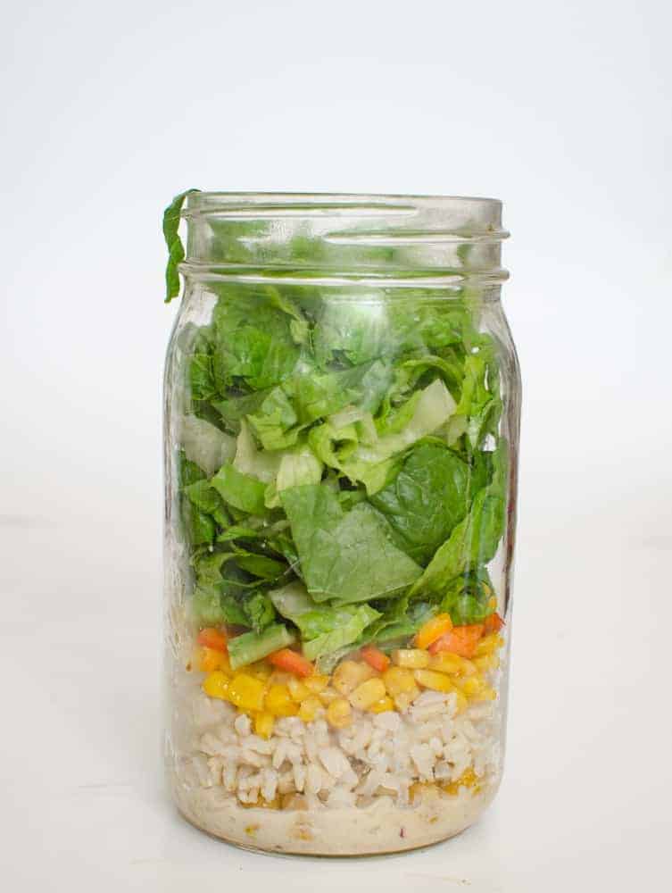 A glass mason jar filled with layered salad ingredients—rice at the bottom, followed by corn and diced carrots, reminiscent of chicken noodle soup flavors, topped with chopped green lettuce. The jar sits on a plain white surface.