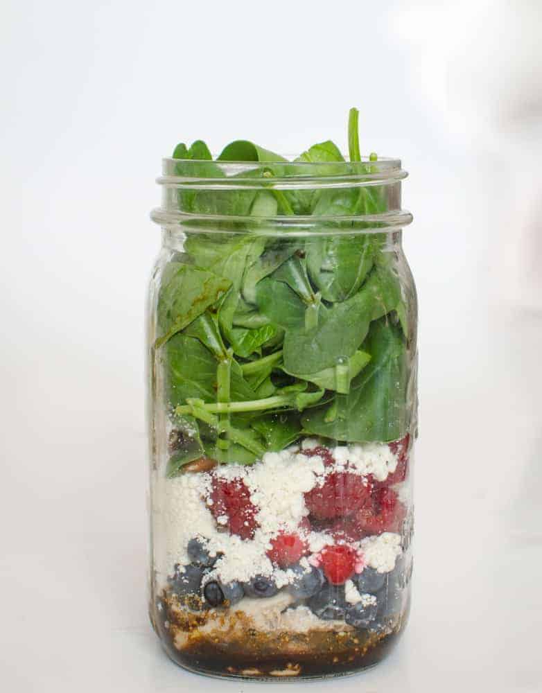 A mason jar filled with layers of salad ingredients—spinach leaves, crumbled cheese, berries, and chicken noodle soup-inspired dressing at the bottom—sits against a plain white background.