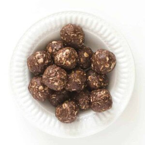 A white bowl filled with round, chocolate peanut butter bites mixed with oats rests on a white background, offering a delectable energy boost.