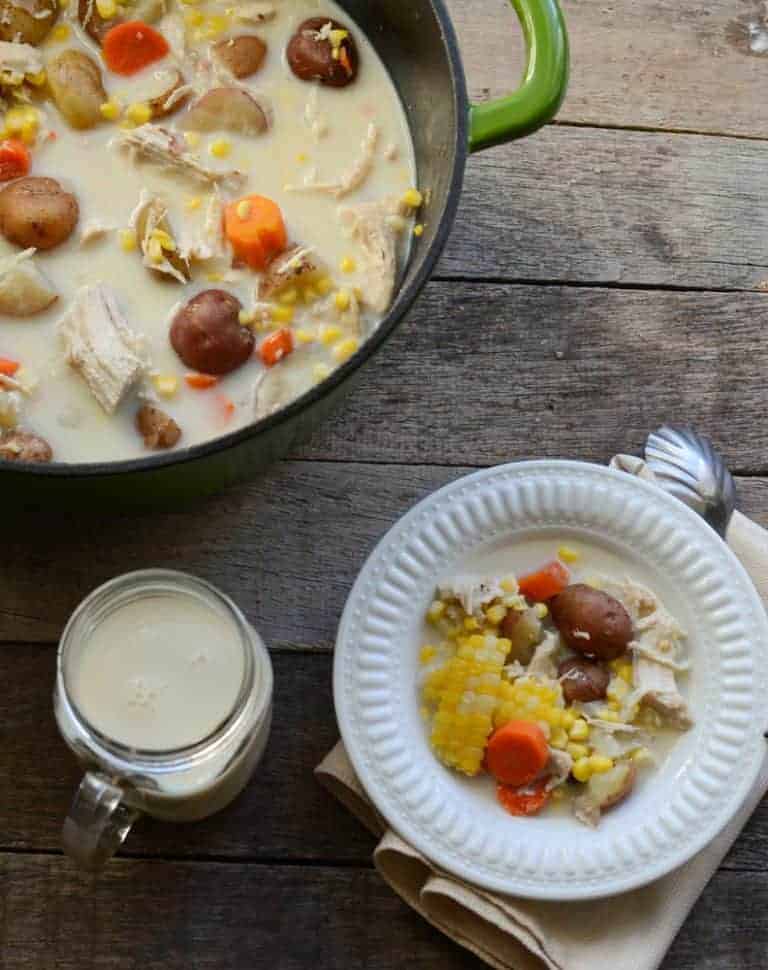 A dutch oven of creamy potato and corn chowder with chicken and carrots and a white bowl with a serving of chowder