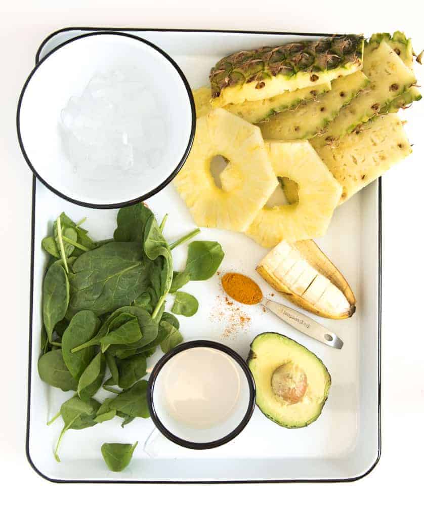 The best workout recovery smoothie to help reduce inflammation, build muscle, and feel your best! And the best part? This recovery smoothie is delicious! The ingredients for a workout recovery smoothie on a white enamelware pan with a black rim, including a bowl of ice, a pile of fresh baby spinach, a couple of pineapple rings, half of a banana, half an avocado, a measuring spoon with ground tumeric, and a white cup with almond milk.