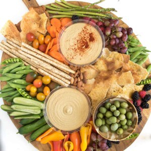 A round wooden hummus platter features two bowls of hummus—one topped with paprika—surrounded by breadsticks, crackers, olives, grape tomatoes, cucumber slices, snap peas, grapes, berries, nuts, mini bell peppers, carrots, and asparagus.