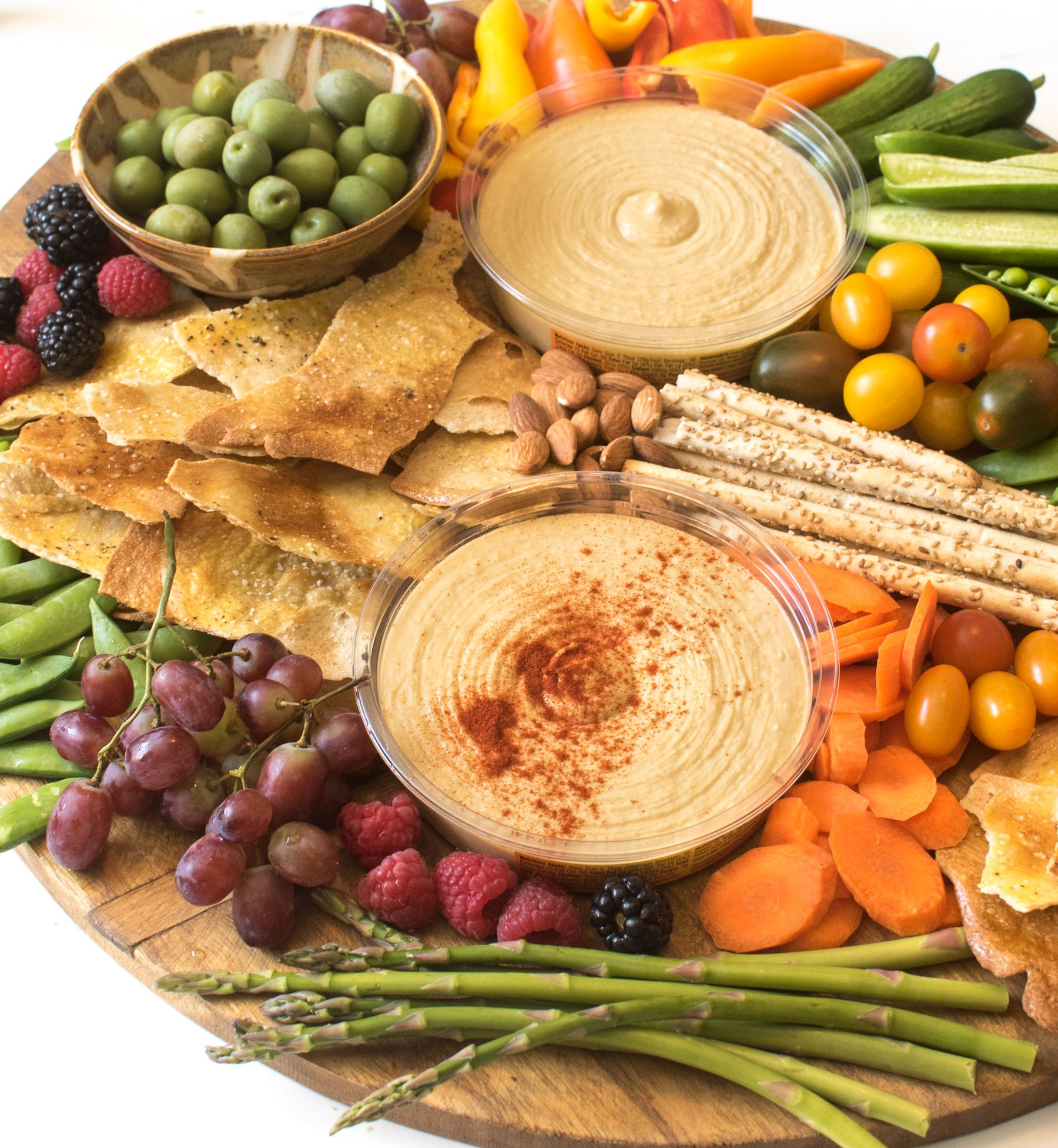 A simple DIY hummus platter is a beautiful addition to any party, or it can be a meal all on it's own! Top view of a Simple DIY Hummus Platter on a round wooden cutting board with a handle that includes two containers of hummus, multiple types of crackers, grape tomatoes, pea pods, berries, asparagus, mini bell peppers, grapes, olives, and almonds.
