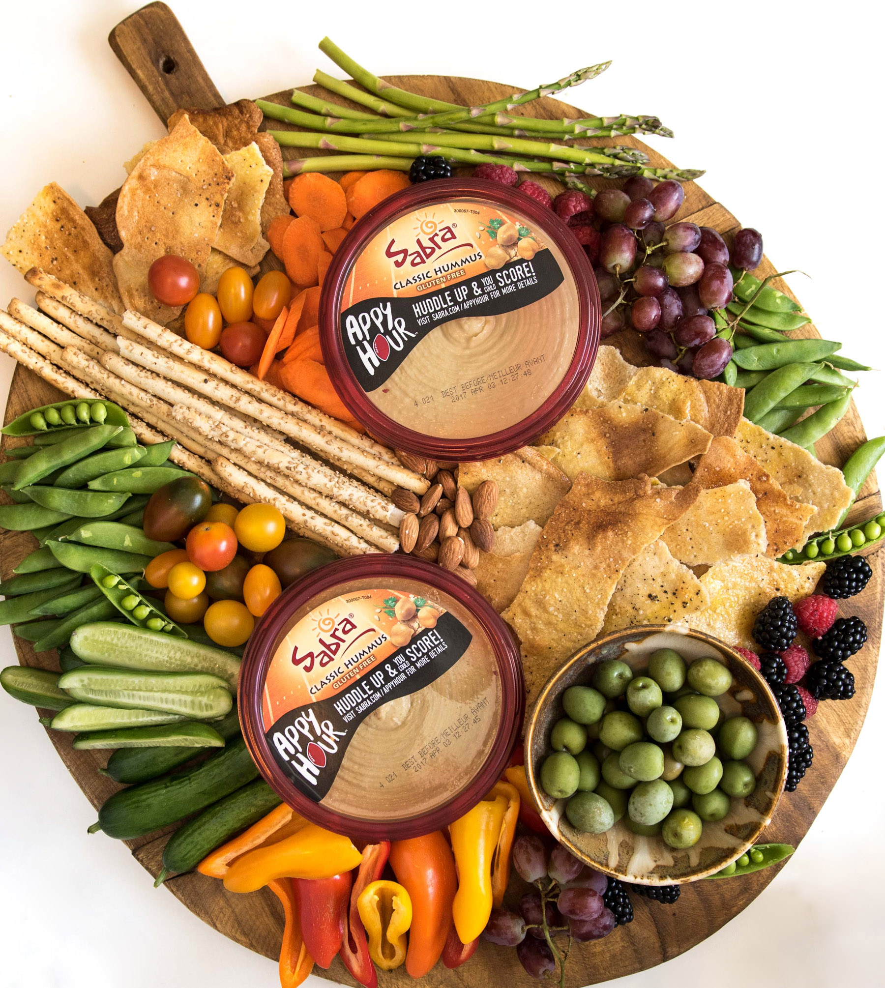 Hummus Platter Top view of a Simple DIY Hummus Platter on a round wooden cutting board with a handle that includes two containers of hummus, multiple types of crackers, grape tomatoes, pea pods, berries, asparagus, mini bell peppers, grapes, olives, and almonds.